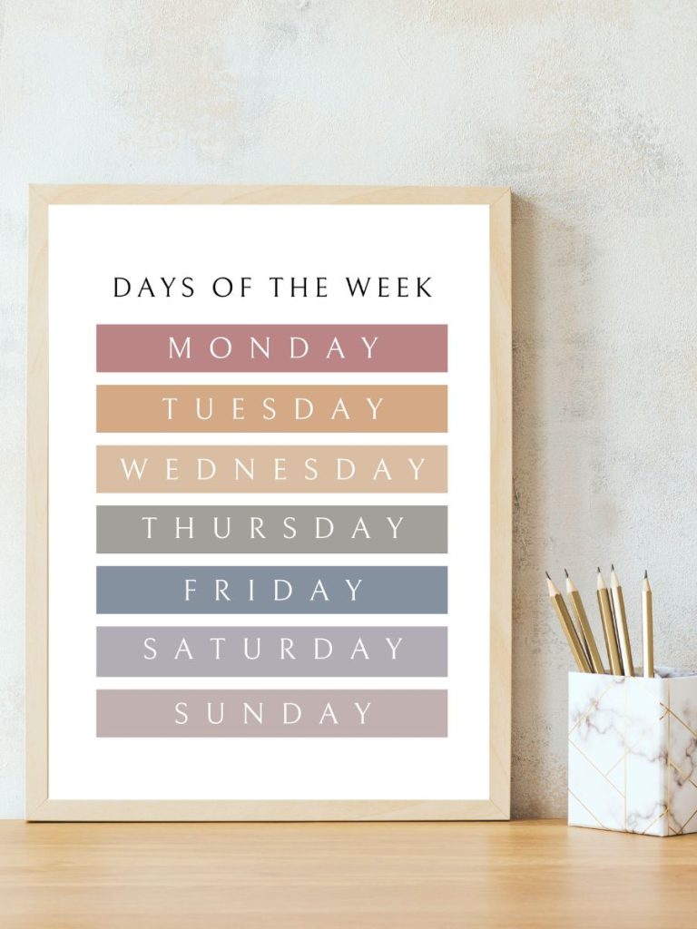 Printable days of the week pastel rainbow color poster in boho wooden frame on desk with pencils 
